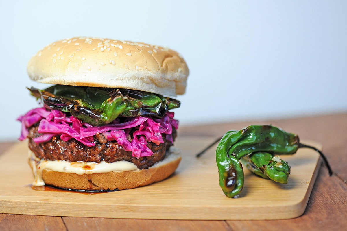 teriyaki burger on wood platter topped with red cabbage and shishito peppers.