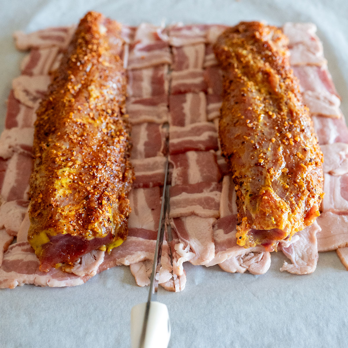 two pork tenderloins on bacon weave while knife is slicing weave in half.