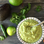 silver bowl of salsa surrounded by avocado, jalapeno and herbs on black board