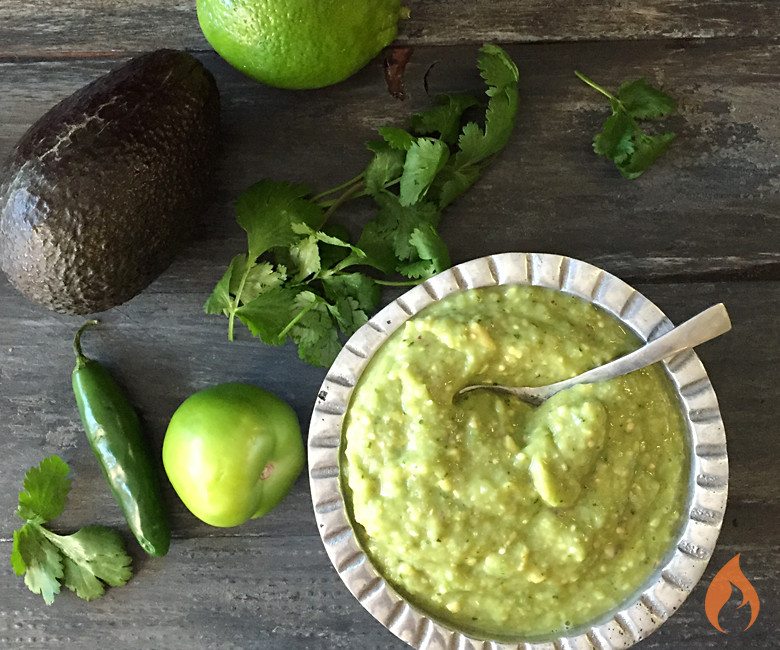 silver bowl of salsa surrounded by avocado, jalapeno and herbs on black board