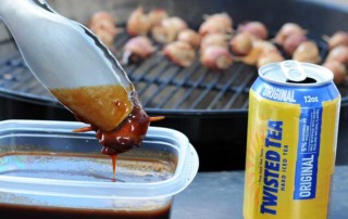 tongs dipping meatball in bbq sauce with grill in background and can of Twisted Tea to right