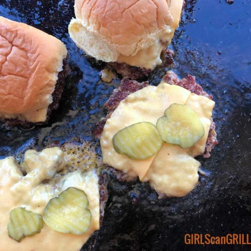 two burger patties on a griddle with cheese and pickles and two buns