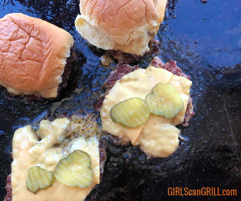 two burger patties on a griddle with cheese and pickles and two buns