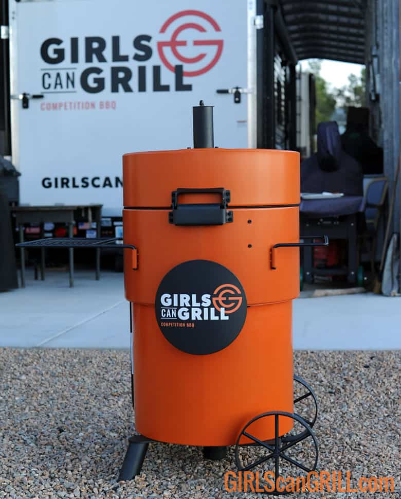 orange Oklahoma Joe's Bronco Pro Drum Smoker with Girls Can Grill logo plate near branded trailer.
