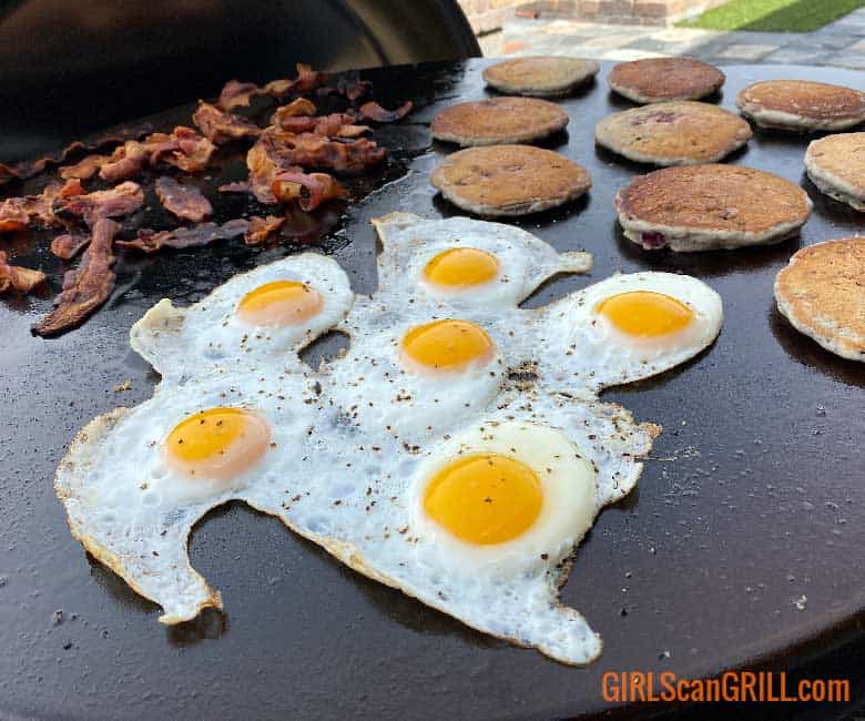 six fried eggs on a griddle with pancakes and bacon