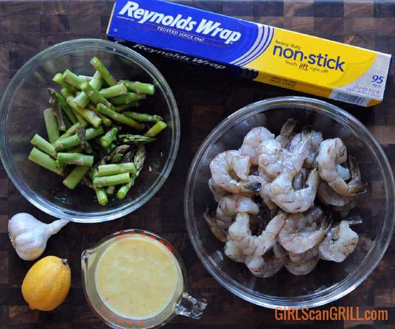 bowl of asparagus, shrimp and lemon garlic sauce with box of Reynolds Wrap® Non-Stick Aluminum Foil box in front of grill with sheets of foil on grates