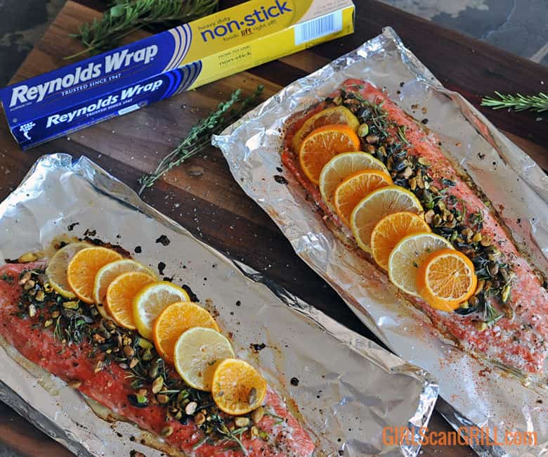 two salmon fillets topped with citrus, herbs and nuts on foil.