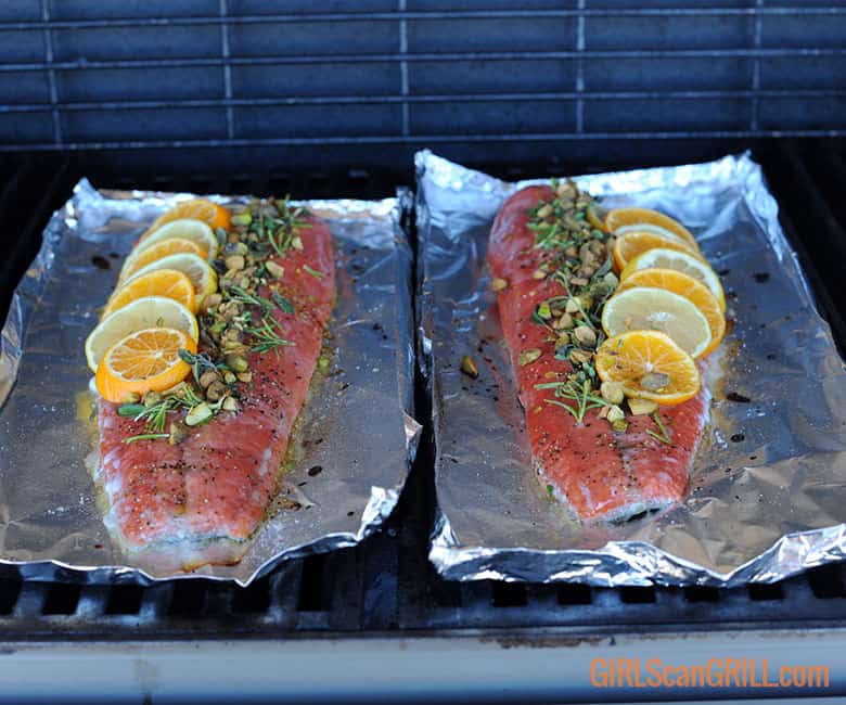 two salmon fillets topped with citrus, herbs and nuts on foil on grill.
