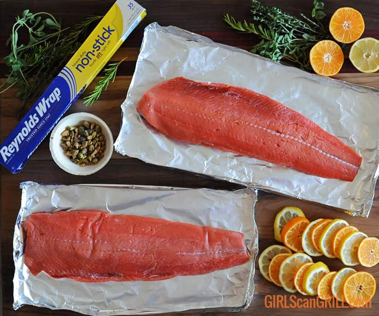 two salmon fillets on foil with citrus slices, herbs and nuts on side.