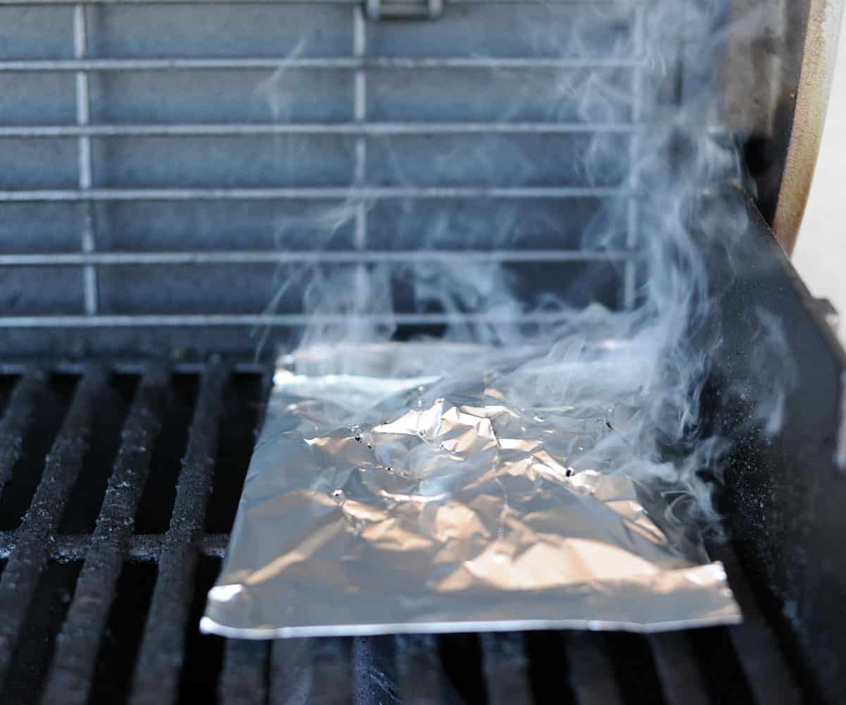 A foil pouch full of wood pellets smoking on a gas grill.