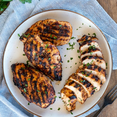 four grilled chicken breasts on a white plate, one sliced.