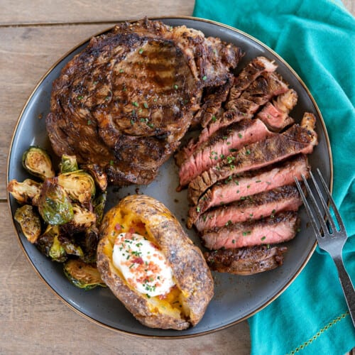 plate of grilled ribeye steak on gray plate with baked potato and grilled brussels sprouts.