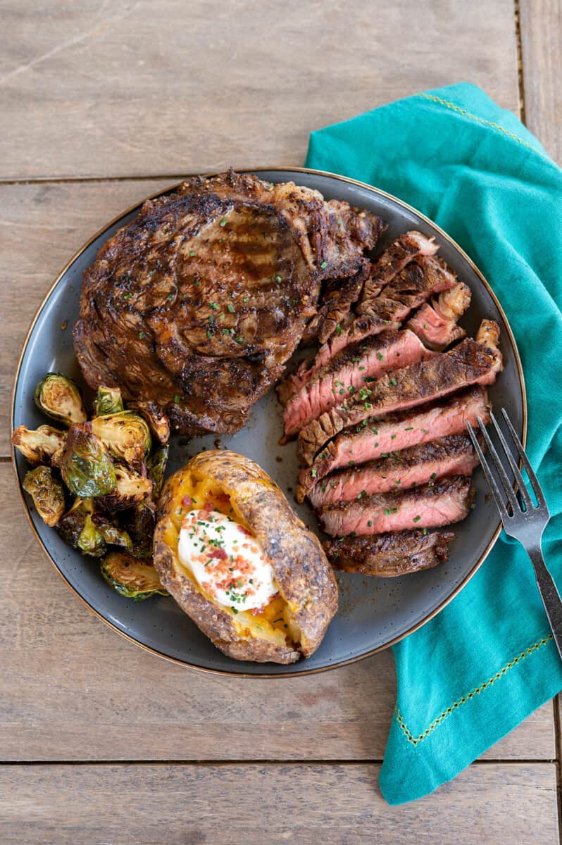 plate of grilled ribeye steak on gray plate with baked potato and grilled brussels sprouts.