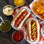 three hot dogs with toppings near bowl of chips.