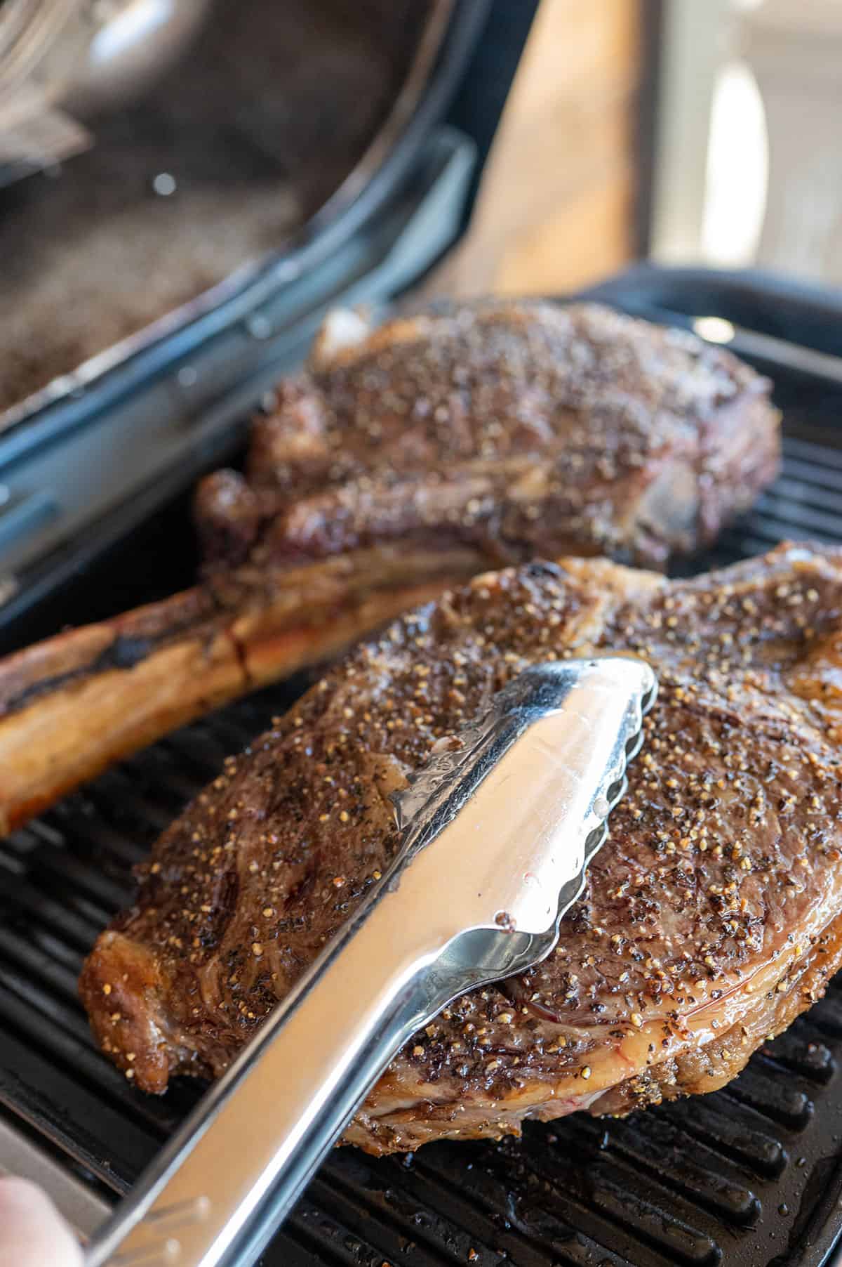 tongs pressing ribeye steak onto grill to form grill marks.