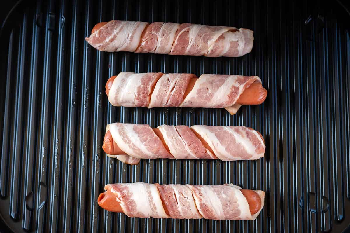 4 bacon-wrapped hot dogs raw on a grill.