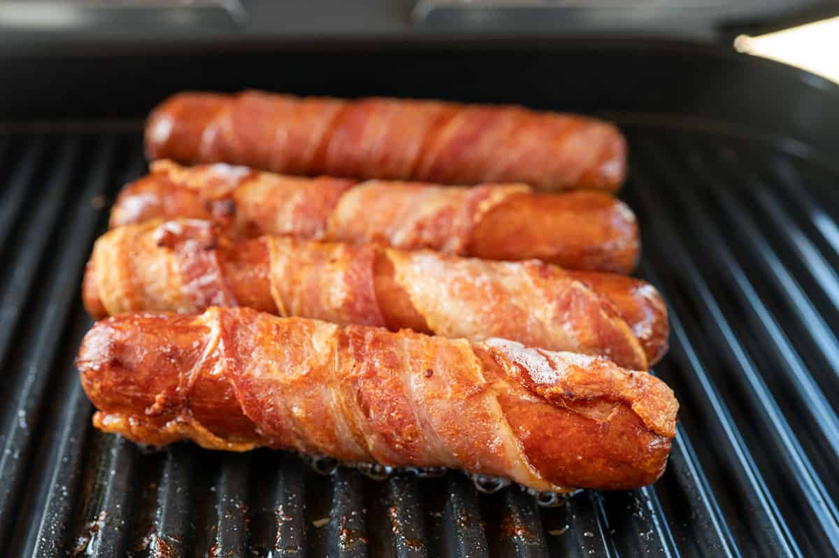four bacon-wrapped hot dogs crisped up on a grill.
