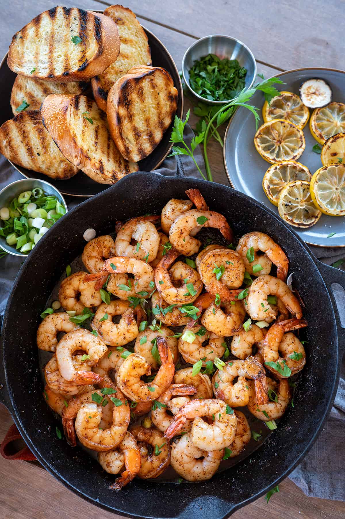 Cast iron skillet full of BBQ shrimp next to grilled bread and grilled lemon slices.