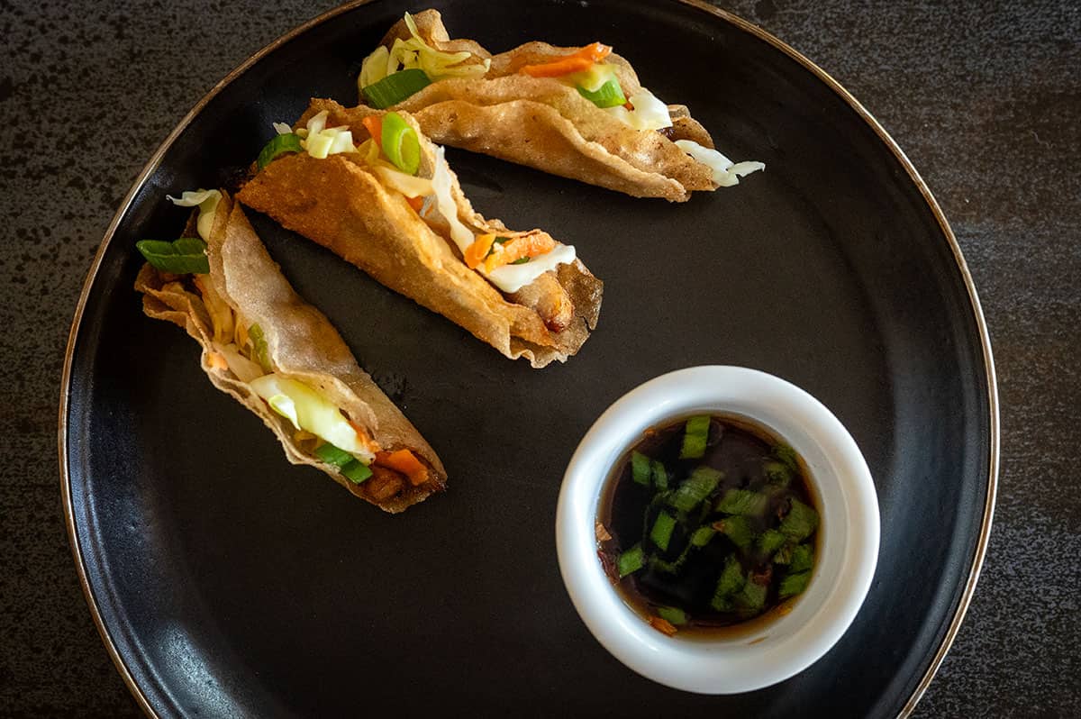 Three chicken lumpia tacos on a plate with dipping sauce.