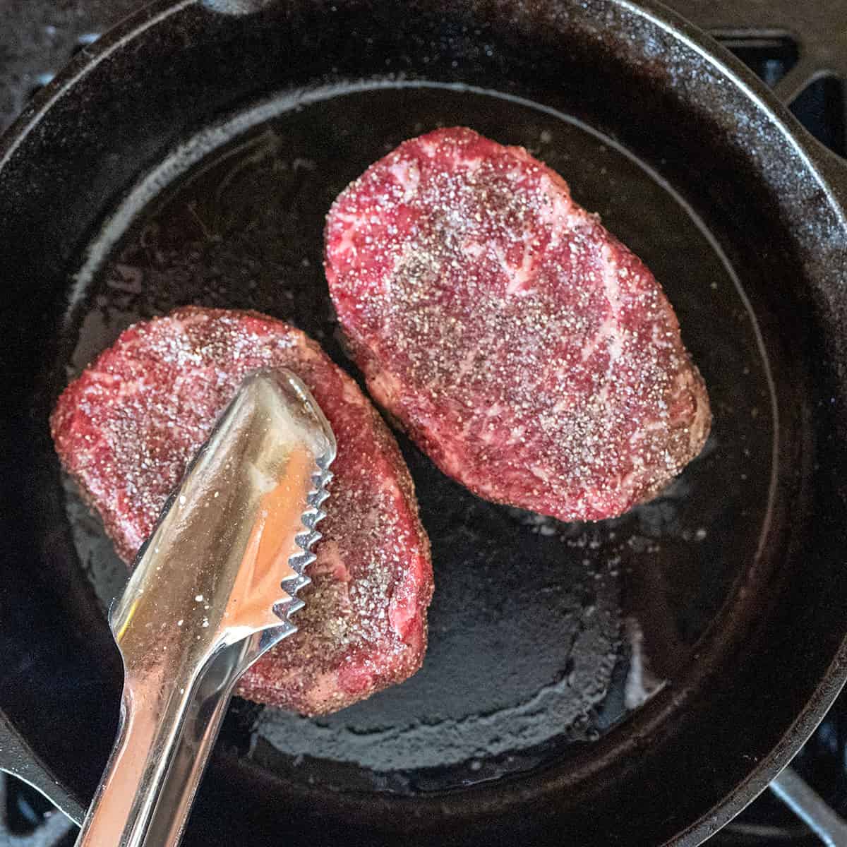 Pressing raw steak down in pan with tongs.