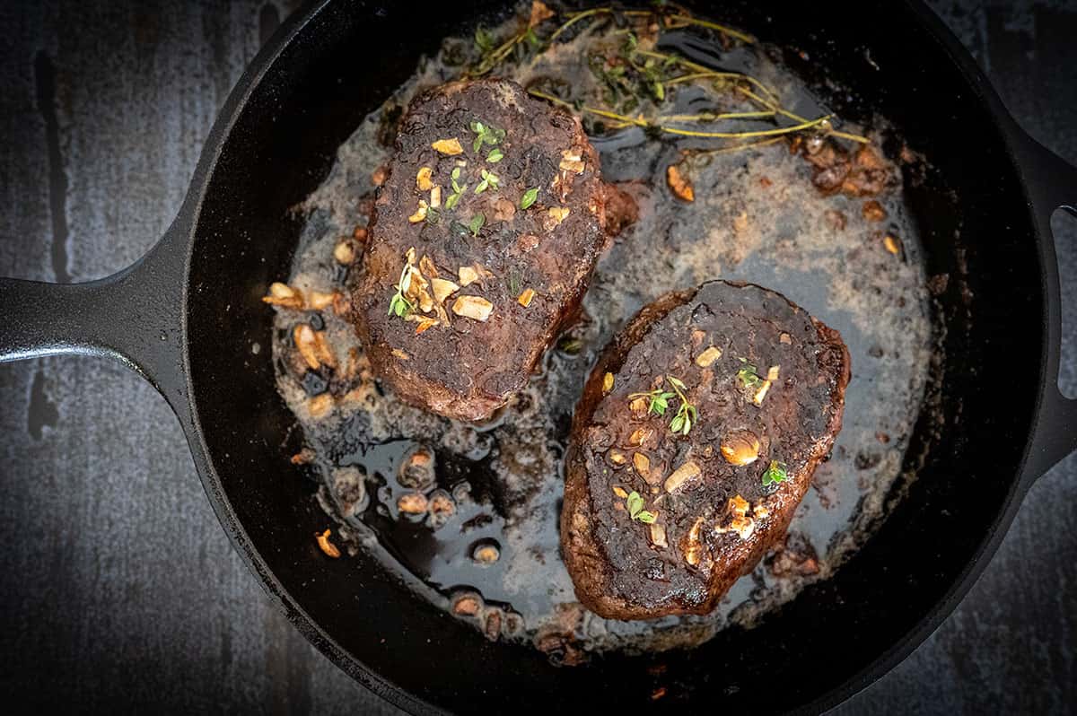 Two whole steaks in cast iron skillet with herb butter.