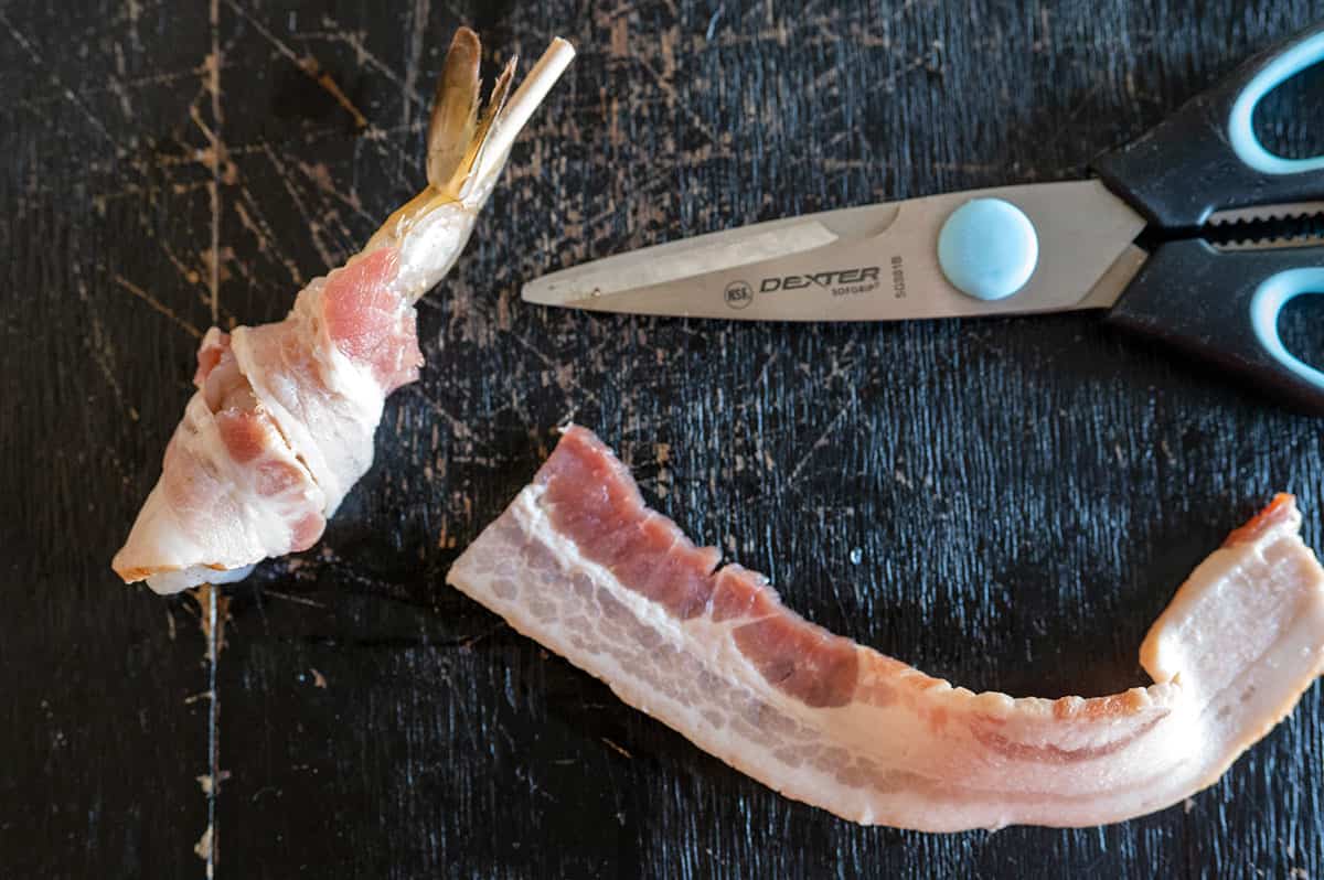 Half slice of bacon wrapped around shrimp with the other half on a board next to a pair of scissors.