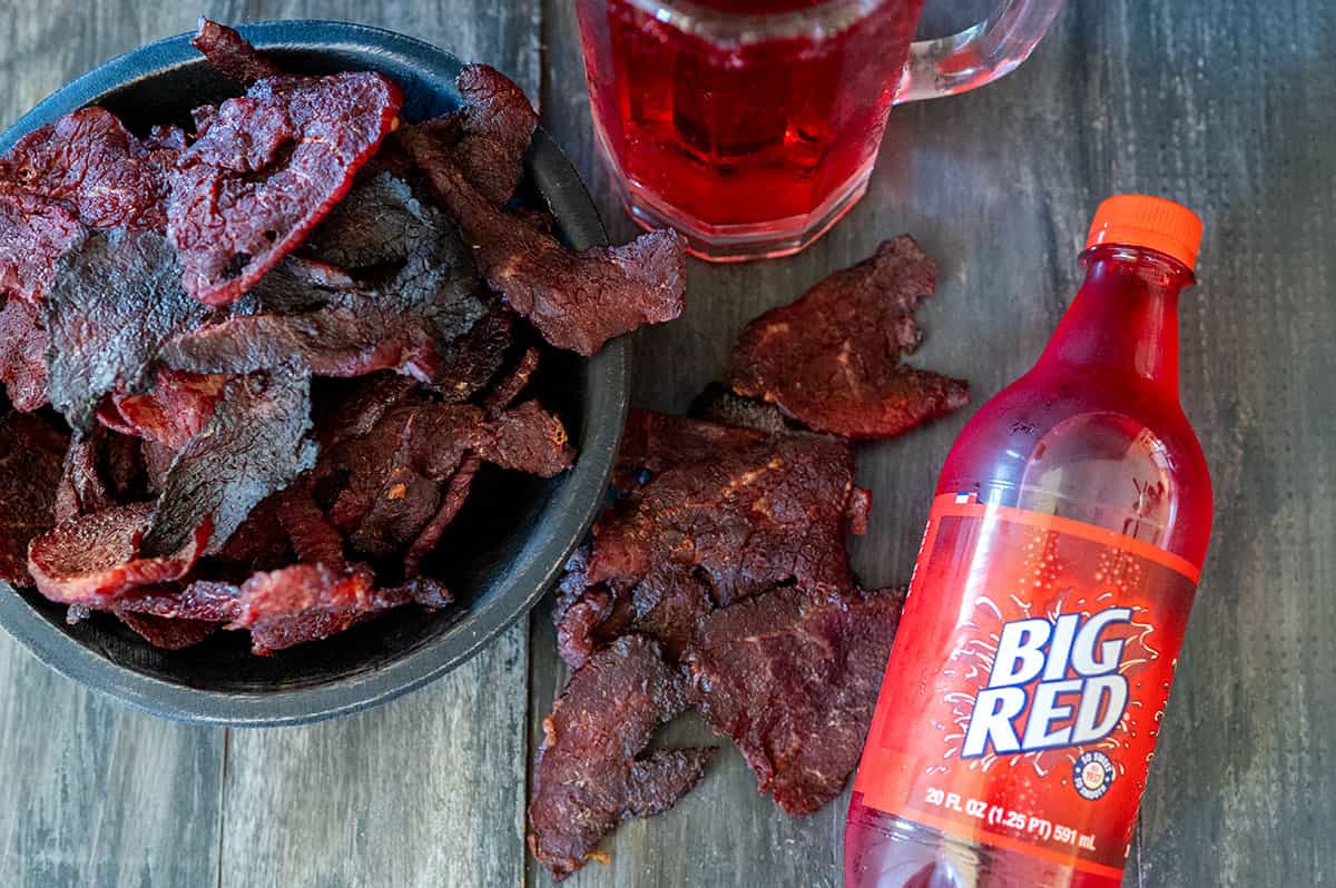 Bowl of Big Red beef jerky next to bottle and mug of Big Red soda.