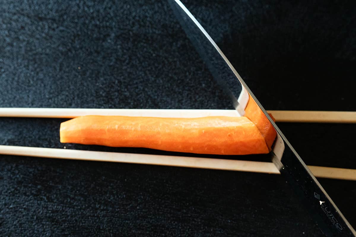 Carrot on cutting board between two chopsticks. Slicing at 45-degree angle.
